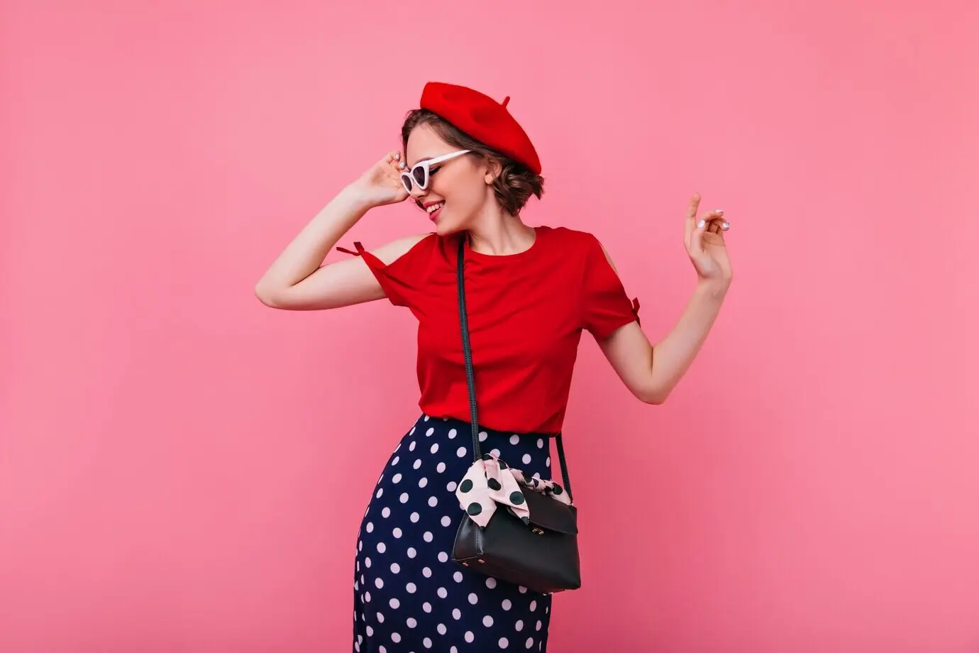 Hübsche, trendige Frau mit Barett, die ihre Sonnenbrille berührt. Innenaufnahme einer fröhlichen, lockigen, jungen Brünette in französischem Outfit.