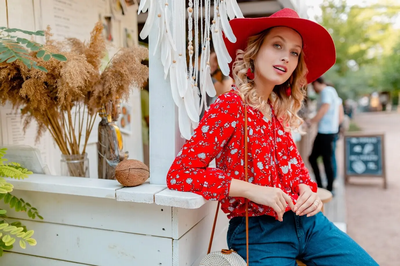 Attraktive, stilvolle, blonde, lächelnde Frau in einem Sommermode-Outfit mit rotem Strohhut und Bluse im Café.